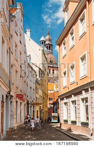 Riga, Latvia - July 2, 2016: People Walking On Narrow Cobbled Kramu Street Of Old Town With View Of Ancient Architectural Landmark Riga Dome Cathedral Under Summer Blue Sky
