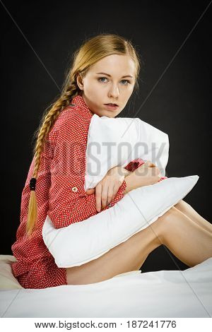 Mental health depression insomia concept. Sad depressive young woman teen blonde girl wearing red pajamas sitting on bed embracing pillow on dark black night background
