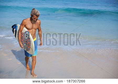 Full-length portrait of an attractive bearded curly man with a surfboard is walking on a beach located on the left side of the frame