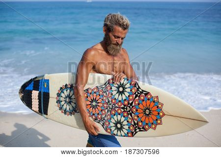 Half-length portrait of an attractive bearded curly man with a surfboard is walking on beach