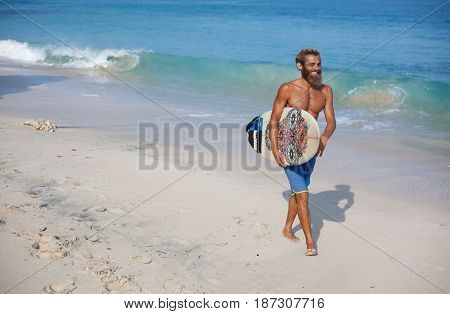 Full-length portrait of an attractive bearded curly man with a surfboard is walking on a beach located on the right side of the frame
