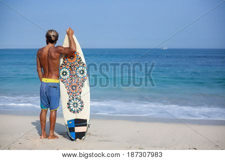 Attractive bearded curly man with a surfboard is standing with his back on a beach and looking to the ocean located on the left side of the frame