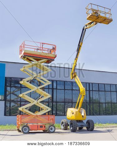 Wheeled scissor lift and wheeled articulated lift with telescoping boom and basket on an asphalt ground against the sky and an industrial building