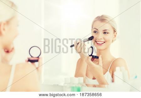 beauty, make up, cosmetics, morning and people concept - smiling young woman applying blush with makeup brush and looking to mirror at home bathroom