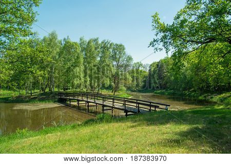 The photo shows a small bridge, a wooden footbridge connecting the island's overgrown island. The footbridge runs over the surface of the lake. It is sunny day.