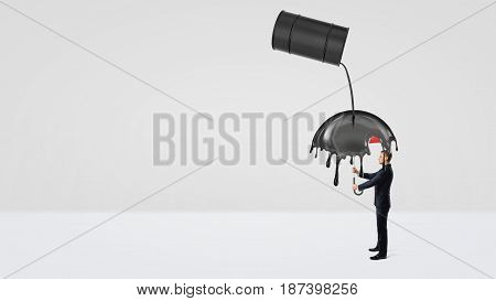 A small businessman hiding under an umbrella from crude oil leaking of a large black barrel. Oil and gas industry. Counting costs and losses. Safety and protection.