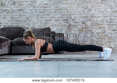 Side view of slim female athlete standing in plank position on floor strengthening core muscles indoors.