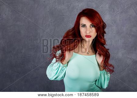 Portrait of beautiful redhead young woman with big boobs in a turquoise dress on a gray background.