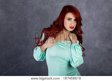 Portrait of beautiful redhead young woman with big boobs in a turquoise dress on a gray background.