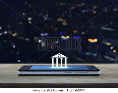 Bank icon on modern smart phone screen on wooden table over blur colorful night light city tower Mobile banking concept