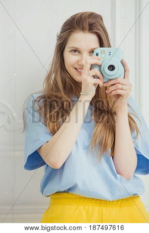 Smiling girl in casual cloth makes photo by portative camera on background of wall. Photographer young woman with long hair dressed in cosy home clothes posing in blue t-shirt and yellow trousers,