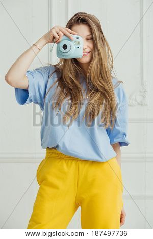Smiling girl in casual cloth makes photo by portative camera on background of wall. Photographer young woman with long hair dressed in cosy home clothes posing in blue t-shirt and yellow trousers,