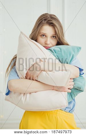 Just woken up girl without make up stands with pillows in hands. Sleepy lady with long luxury hair in the morning. Drowsy woman hugging big pillow