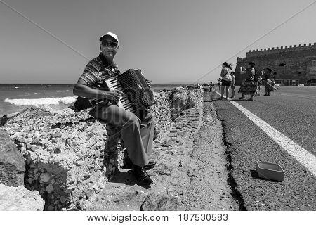 HERAKLION GREECE - JULY 16 2016: Crete. Musician on the waterfront plays accordion. In the background Venetian fortress. Black and white.