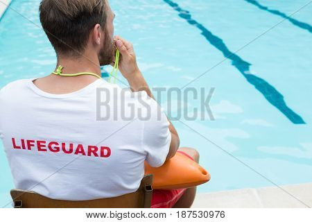 Rear view of lifeguard sitting on chair and blowing whistle at poolside