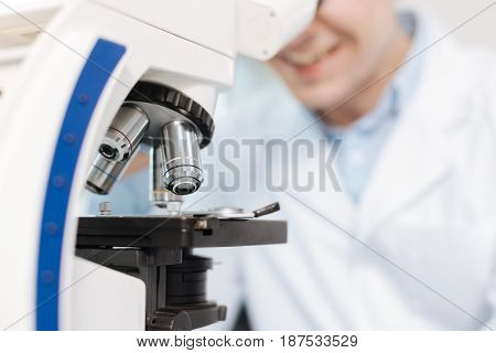 Check details. Smiling practitioner using microscope for studding genes sitting at his workplace while looking into ocular