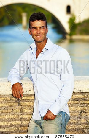 Beautiful smiling man outdoors. Tiber river from the bridge. A beautiful young Italian man posing outdoors on Milvian Bridge in the historic center of Rome, Italy. Blue shirt and jeans.