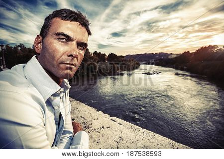 Lonely man looking suspicious. River at sunset. Italian handsome man on the bridge looking serious and confident attitude. Tiber river and the evening sky at sunset. Rome, Italy.