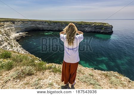 girl in white t-short with hands up standing on a cliff near sea bay