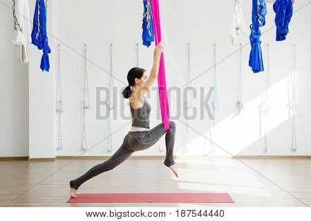 Girl stretching legs with help of hammock. Aerial exercise or antigravity yoga indoors, woman jumps and does aero poses with head leaned