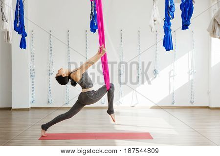 Girl stretching legs with help of hammock. Aerial exercise or antigravity yoga indoors, woman jumps and does aero poses with head leaned to back