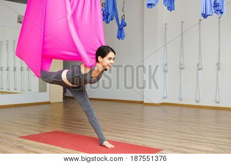 Girl stretching leg with help of hammock. Aerial exercise or antigravity yoga indoors, woman does aero poses with head leaned to back