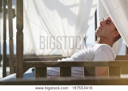 Relax serenity at the beach. Man lying on canopy wooden beds white curtains. Closed eyes and contemplation. Holiday concept, serenity and peace of mind. Intense and warm lights