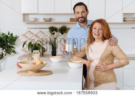 Expecting husband embracing his riant pregnant wife while both posing and looking at camera in the kitchen.