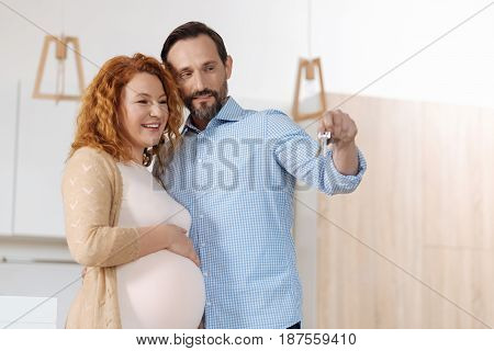 We are moved. Smiling married couple holding keys to their new house, husband hugging wifes waist while both huddling up to one another.