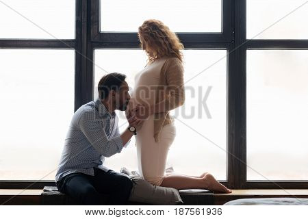 Tender sentiment. Mature expectant mother standing on her knees on a pillow and watching her husband kissing a pregnant belly.