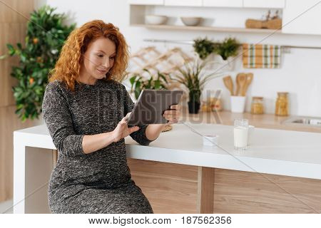 Browsing the net. Full term pregnant woman wearing dress sitting in the kitchen and using a tablet while having a lunch at home.