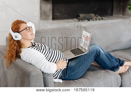 Relaxation and calm. Beautiful preggy woman lying on a sofa with a laptop on her legs and listening to classical song while having a rest at home.
