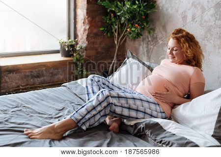 Cant wait to see you. Happy mother-to-be relaxing at home, while lying on a bed and looking on her baby moves.