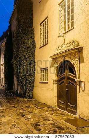 Riga, Latvia. Old Wooden Brown Door Of Portal, Decorated By Molding On Facade Of Ancient Building On Torna Street In Old Town In Lighting At Evening Or Night Illumination In Old Town. Blue Hour