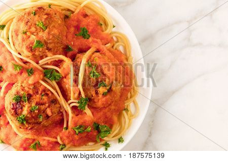 A closeup of a plate of meatballs with pasta, tomato sauce, and fresh parsley, shot from above on a white marble texture with a place for text