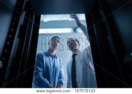 Experienced technicians. Joyful positive nice men standing behind the metal grid of the data server and smiling while working as technicians in the data center