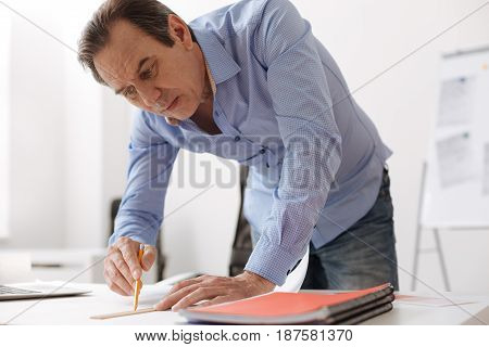 Do it in such a way. Professional serious senior engineer drawing a blueprint and stnding near table in the office