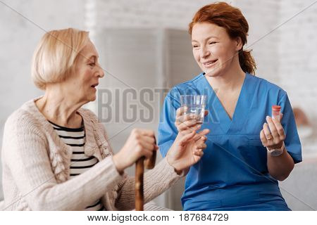 Time to get healthy. Cheerful focused skillful woman giving her patient a glass of water for taking her pills while taking care of old ladies health