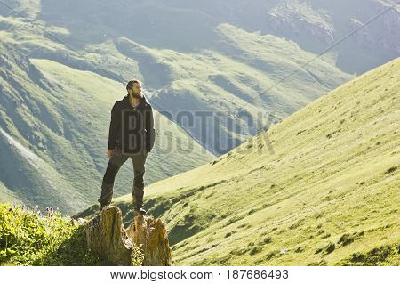 man with beard in black jacket standing with hands up above mountain slopes at yellow sunset