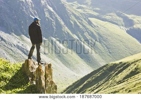 man in black jacket standing with hands up above mountain slopes at yellow sunset