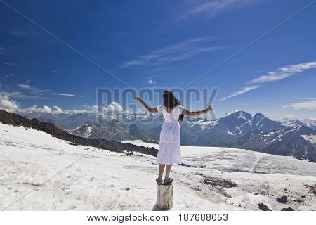 girl in white dress standing on snow slope of caucasus mountains with hands-up with mountains surrounding and blue sky