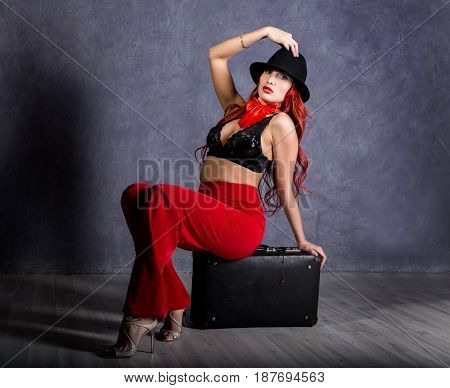 woman in red pants and black hat with big boobs sits on a retro suitcase.