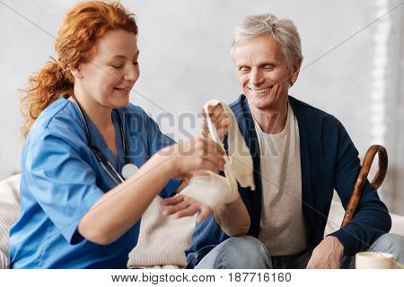 Protecting the joint. Distinguished positive medical worker treating her elderly patient whom she visiting at home by applying special medical bandage