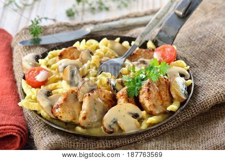 Fried medallions of pork fillet with Swabian spaetzle (so called Schwabenpfandl or Schwabentopf ) and mushroom cream sauce served in an iron frying pan on a rustic table with an old jute sack