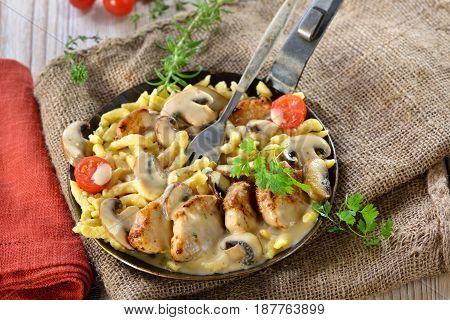Fried medallions of pork fillet with Swabian spaetzle (so called Schwabenpfandl or Schwabentopf ) and mushroom cream sauce served in an iron frying pan on a rustic table with an old jute sack
