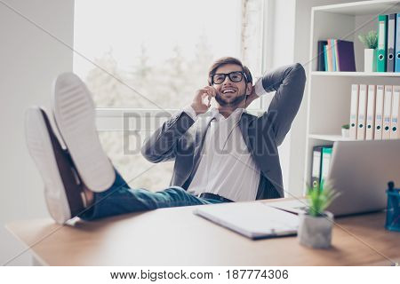 Cheerful Young Successful Man Is Talking On The Phone At The Workplace With Feet On The Table. He Is