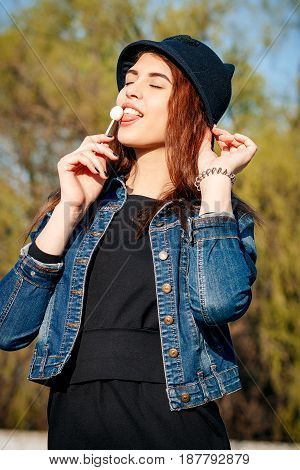 Cheerful Stylish Young Girl In Hat, And Jeans Jacket With Candy On Stick, Chupa Chups, On Sunny Day