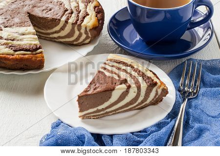 Delicious homemade chocolate cake with cottage cheese Zebra (Marble cake). Tasty breakfast. Piece of cake on blue plate blue cup and blue gauze textiles on white wooden table. Selective focus