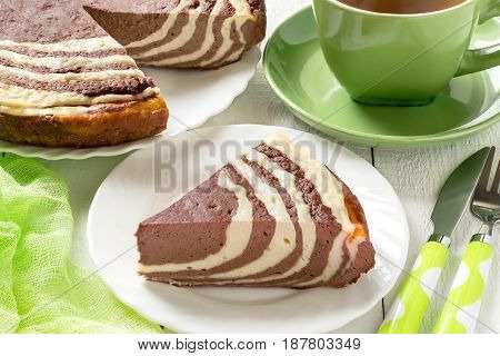Delicious homemade chocolate cake with cottage cheese Zebra (Marble cake). Tasty breakfast. Piece is cut from cake green cup of tea and green gauze textiles on white wooden table. Selective focus