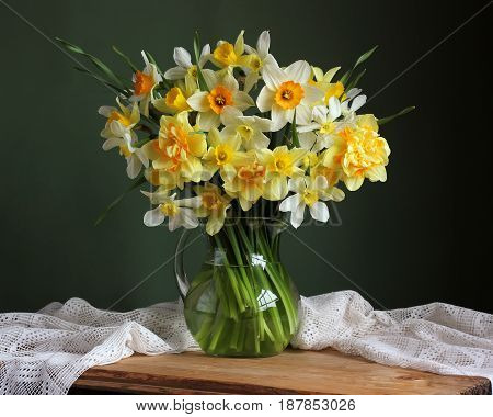 Varietal yellow daffodils in a jug on table with white tablecloth against a green background.